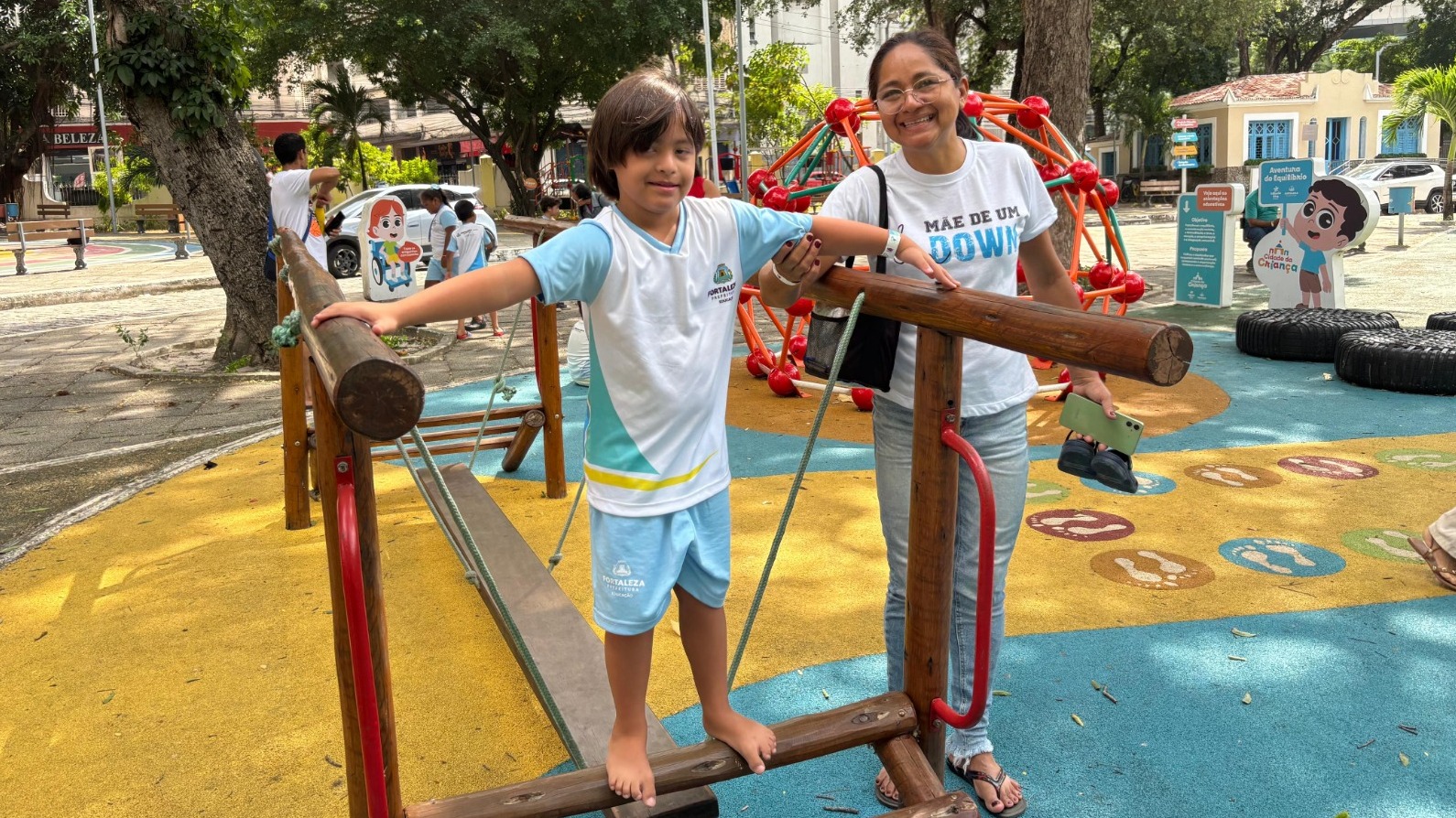 Uma criança e uma mulher adulta posam para a foto na Cidade da Criança. A criança está em cima de um brinquedo de madeira